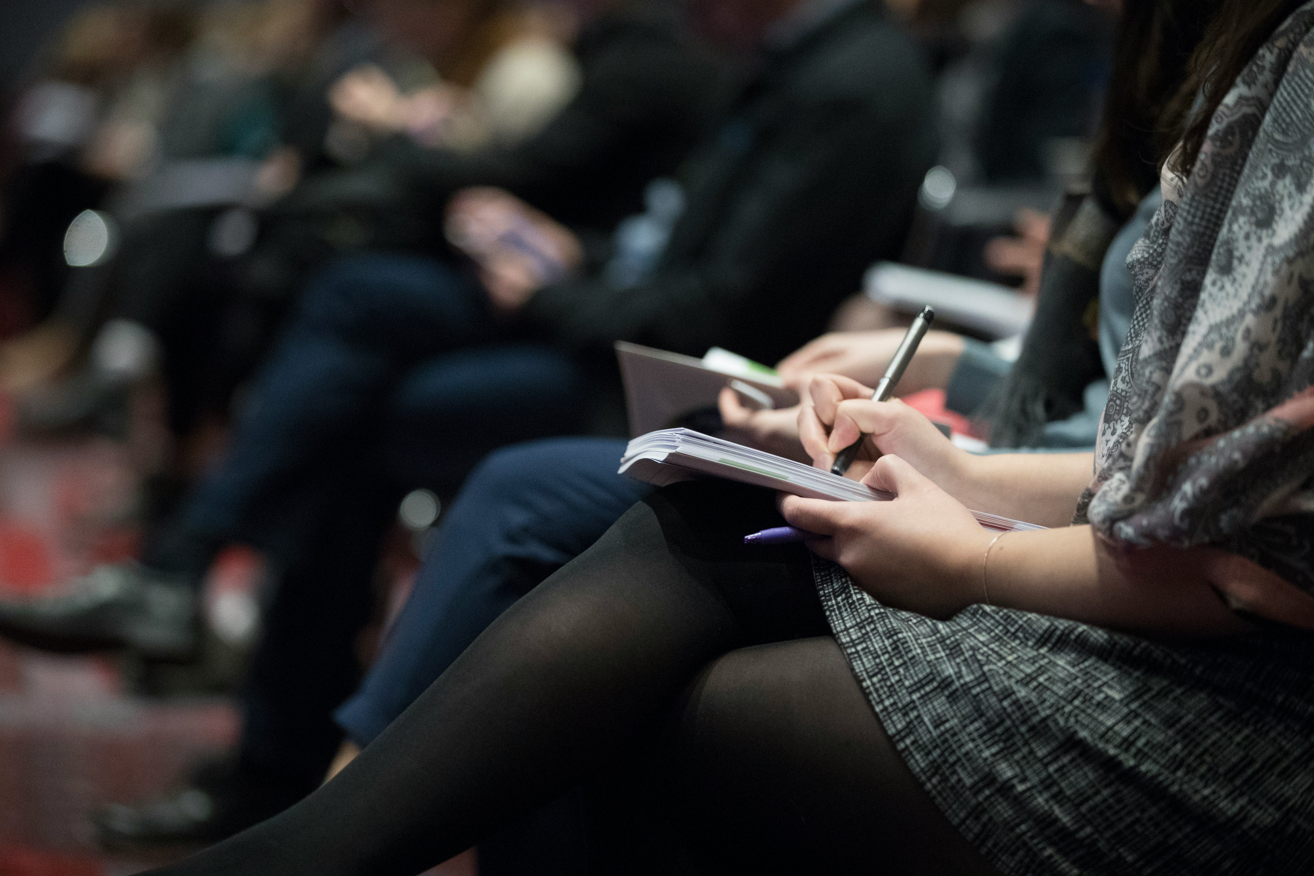 Front row of seated event attendees, writing notes during a workshop presentation.
